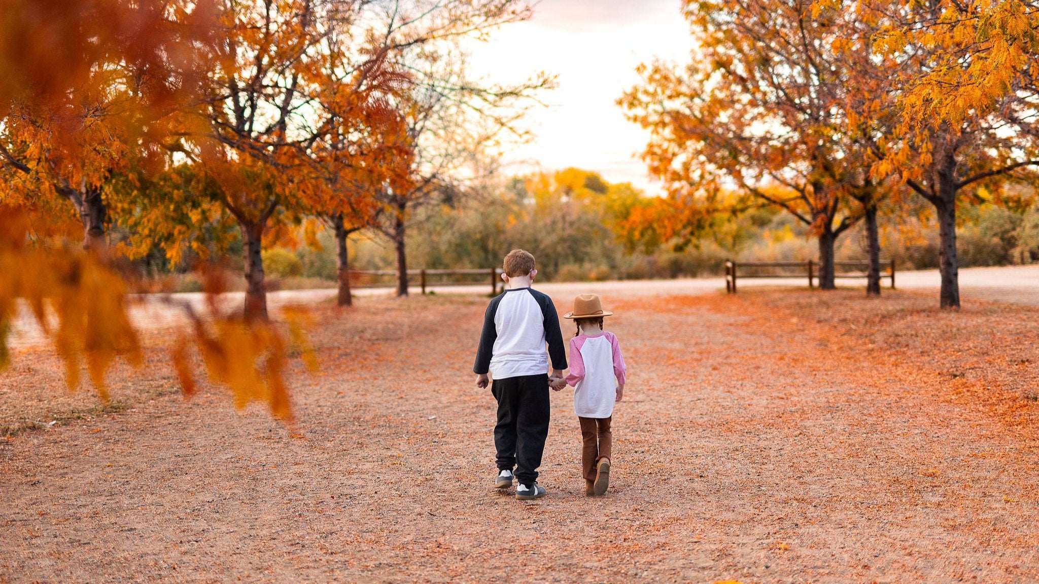 Two children holding hands, walking through a beautiful autumn landscape with vibrant orange leaves, wearing cozy raglan tees from Jumpin Jaks Tees. Perfect fall apparel for kids, blending comfort and style. Ideal for family outings, fall photoshoots, and Thanksgiving gatherings.